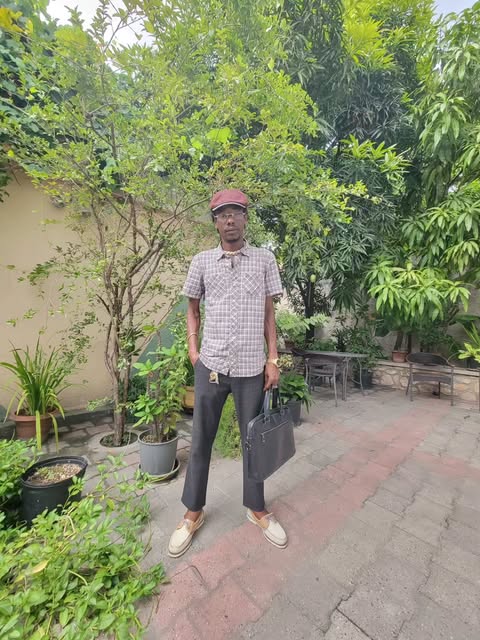 Man in a plaid shirt, cap and grey pants in a leafy courtyard